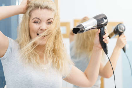 Positive woman using hair dryer on her blonde hairdo. Haircare, hairstyling concept.の写真素材