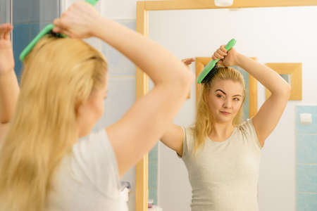 Happy woman combing her hair with brush. Young smiling female with natural blond straight long hairs in bathroom after morning shower.の写真素材