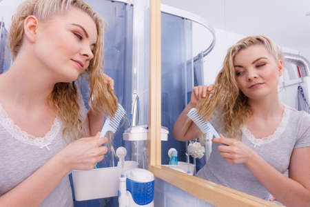 Woman combing after bath her long wet curly hair in bathroom, looking in mirror. Blonde girl taking care refreshing her hairstyle in morning. Haircare concept.の写真素材
