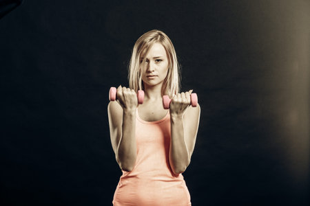 Fitness, sport, training and lifestyle concept. Fit woman with dumbbells flexing muscles in gym. Muscular blonde girl lifting light weights studio shot on black backgroundの写真素材