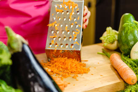 Unrecognizable woman grating carrot on metal grater, kitchen utensil making food preparing healthy vegetable salad.の写真素材