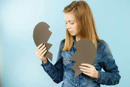 Sad young teenage woman with broken heart made of paper. Negative sad emotions, relationship problems concept. Shot on blue.の写真素材