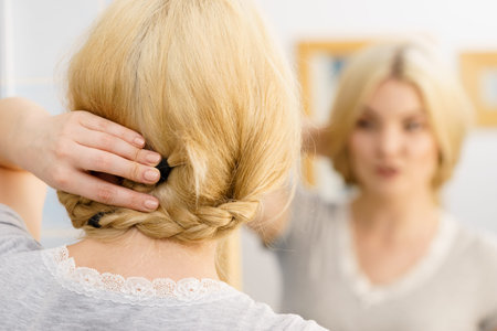 Happy positive blonde woman in bathroom during morning routine looking at herself in mirror styling...