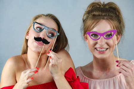 Two happy women holding symbols on stick having fun pretending to wear eyeglasses. Photo and carnival funny accessories concept.の写真素材
