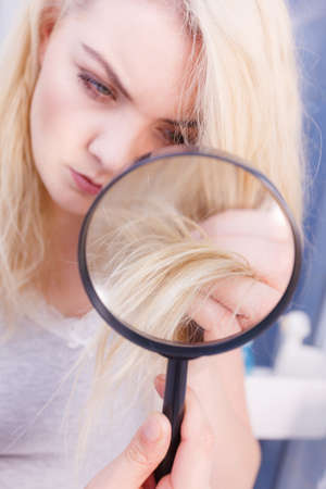 Haircare, bad effects of bleaching concept. Unhappy woman looking at ends of her blonde hair through magnifying glassの写真素材