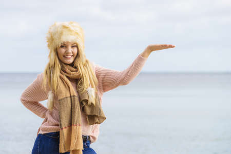 Teenage female spending time outdoor on the beach during autumn weather pointing at copy space. Wearing sweater, scarf and fur hat.の写真素材