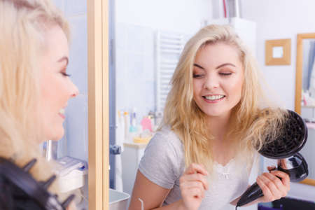 Haircare. Beauty long haired blonde woman drying hair in bathroom. Smiling girl blowing wind on wet head using hairdryer, doing curls with diffuser nozzle.の写真素材