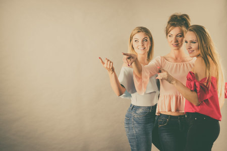 Fashionable adult female friends models pointing at grey copy space. Studio shot