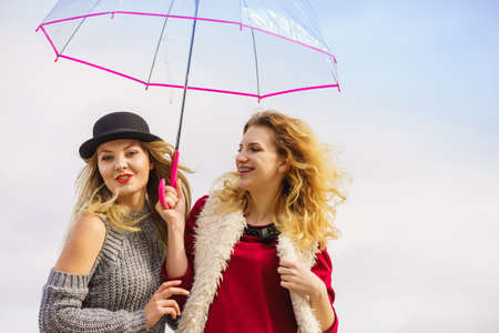 Two fashionable women wearing stylish outfits holding transparent umbrella spending their free time outdoorの写真素材