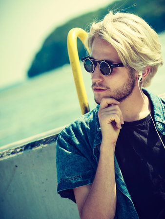 Vacation, relaxation, passion concept. Young fashionable blonde in sunglasses man relaxing, listening to music and enjoying beautiful, sunny weather. Outdoor shotの写真素材