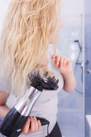 Haircare. Beauty long haired blonde woman drying hair in bathroom. Smiling girl blowing wind on wet head using hairdryer, doing curls with diffuser nozzle.の写真素材