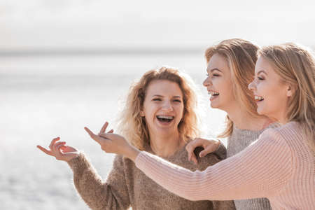 Three fashionable women wearing sweaters during warm autumnal weather spending their free time on sunny beach. Fashion models outdoor having fun.の写真素材