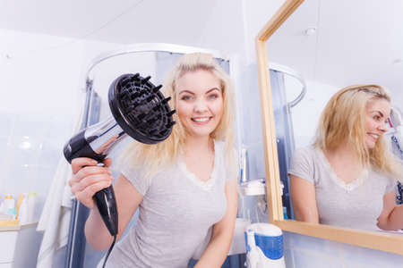 Haircare. Beauty long haired blonde woman drying hair in bathroom. Smiling girl blowing wind on wet head using hairdryer, doing curls with diffuser nozzle.の写真素材