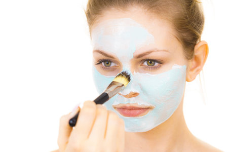 Young woman applying with brush green mud mask to face, on white. Teen girl taking care of oily...