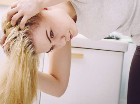 Blonde woman having problems with greasy oily hair in bathroom. Female showing scalp, scratching herself, hairdo loss, dandruff problem.の写真素材