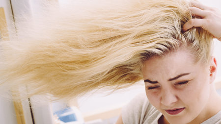 Blonde woman having problems with greasy oily hair in bathroom. Female showing scalp, scratching herself, hairdo loss, dandruff problem.の写真素材