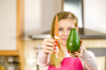 Woman cutting, preparing green vegetable, delicious avocado using kitchen knife. Female wearing...