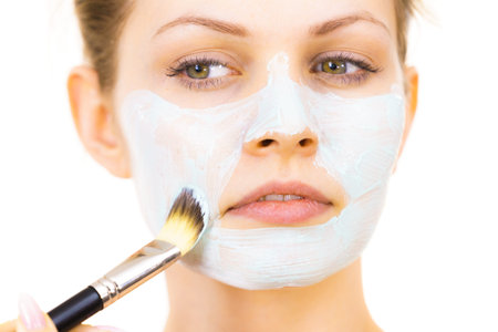 Young woman applying with brush green mud mask to face, on white. Teen girl taking care of oily...