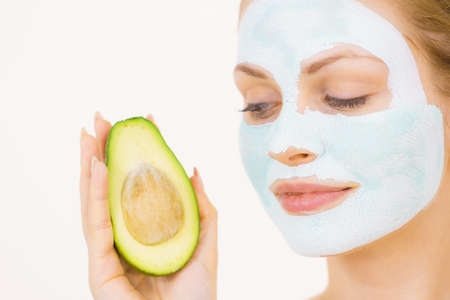Young woman with white green mud mask on face holding avocado fruit. Teen girl taking care of her skin, cleaning the pores. Beauty treatment.の写真素材