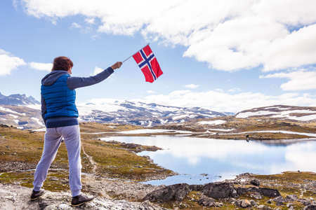 Tourist woman enjoy mountains landscape, holding norwegian flag. National tourist scenic route 55 Sognefjellet, Norwayの写真素材