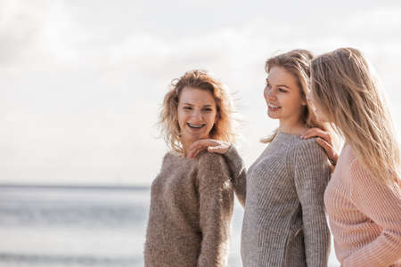 Three fashionable women wearing sweaters during warm autumnal weather spending their free time on sunny beach. Fashion models outdoor having fun.の写真素材