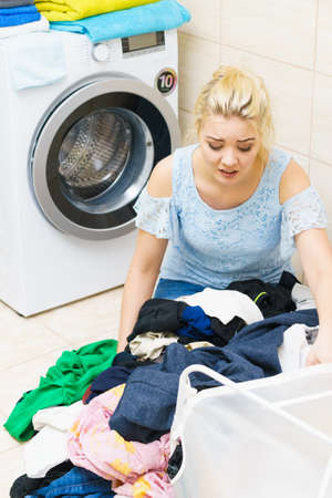 Sad woman having a lot of laundry to make. Unhappy female surrounded by big clothing piles stack sitting next to washing machine being tired houseworking.の写真素材