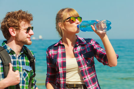 Adventure, summer, tourism active lifestyle. Young couple backpacker tramping by seaside, girl drinking water from plastic bottleの写真素材
