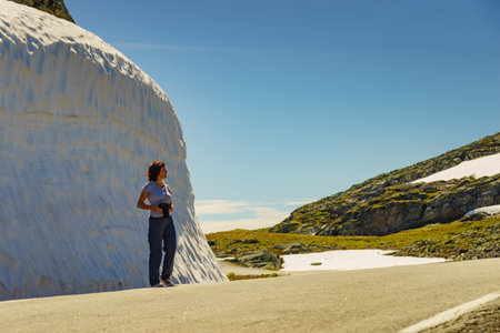 Tourist woman with camera taking travel picture from high snow wall on mountain road Aurlandsfjellet...
