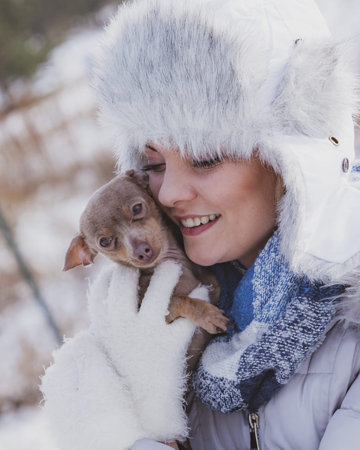Young woman having fun outside in snow. Female playing with her small purebreed dog puppy in cold...