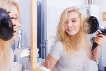 Haircare. Beauty long haired blonde woman drying hair in bathroom. Smiling girl blowing wind on wet head using hairdryer, doing curls with diffuser nozzle.の写真素材
