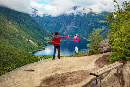 Female tourist enjoying scenic view over fjord Geirangerfjorden from Flydalsjuvet viewpoint, holding Norwegian flag. Cruising vacation and travel.の写真素材