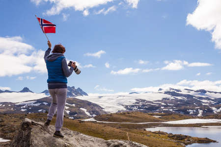 Tourist woman enjoy mountains landscape, holding Norwegian flag and photo camera. National tourist scenic route 55 Sognefjellet, Norwayの写真素材