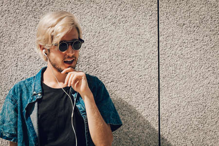 Men fashion, technology, urban style clothing concept. Hipster guy standing on city street wearing jeans outfit and eccentric sunglasses listening to music and holding phoneの写真素材