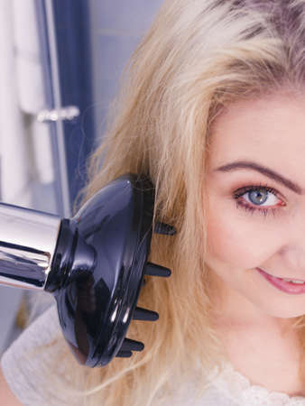Haircare. Beauty long haired blonde woman drying hair in bathroom. Smiling girl blowing wind on wet head using hairdryer, doing curls with diffuser nozzle.の写真素材