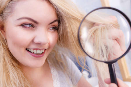 Haircare concept. Happy woman looking at ends of her blonde healthy hair through magnifying glassの写真素材
