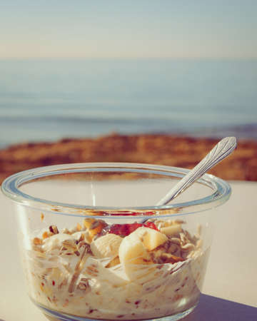 Healthy breakfast on nature. Oatmeal granola with yogurt, banana fruits in bowl, sunny morning, ocean backgroundの写真素材