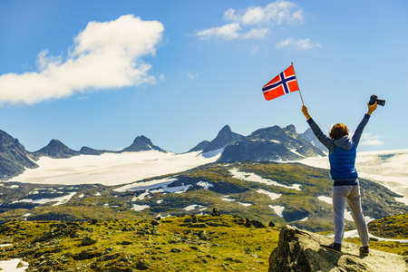 Tourist woman enjoy mountains landscape, holding norwegian flag and photo camera. National tourist scenic route 55 Sognefjellet, Norwayの写真素材