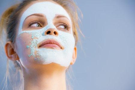 Young woman with white dried mud mask on face being removing cosmetic. Teen girl taking care of oily...