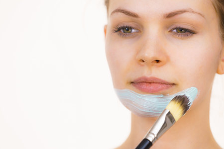 Young woman applying with brush green mud mask to face, on white. Teen girl taking care of oily skin, purifying the pores. Beauty treatment. Skincare.の写真素材