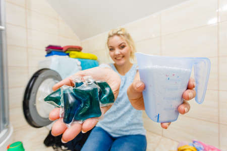 Woman in bathroom doing laundry, choosing the best detergent. Female holding powder container and gel pods for washing clothes. Household duties. Wide angle viewの写真素材