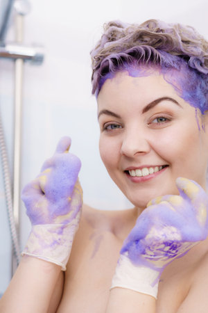 Woman wearing latex protective gloves while applying purple toner shampoo on her blonde hair....