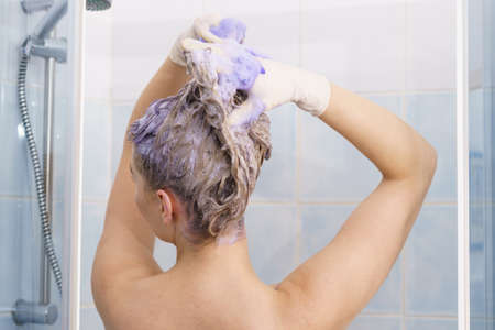 Woman having purple shampoo foam on her head under the shower. Female toning blonde hair using colored shampoo.の写真素材
