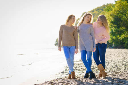 Three fashionable women wearing sweaters during warm autumnal weather spending their free time on sunny beach. Fashion models outdoorの写真素材