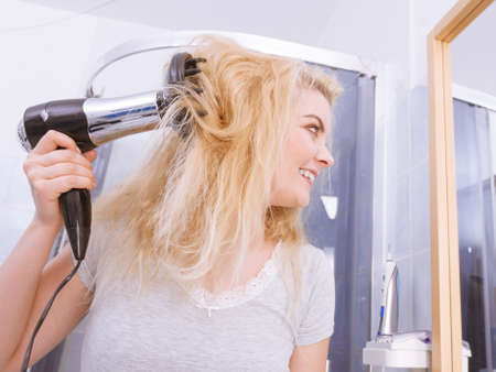 Haircare. Beauty long haired blonde woman drying hair in bathroom. Smiling girl blowing wind on wet head using hairdryer, doing curls with diffuser nozzle.の写真素材