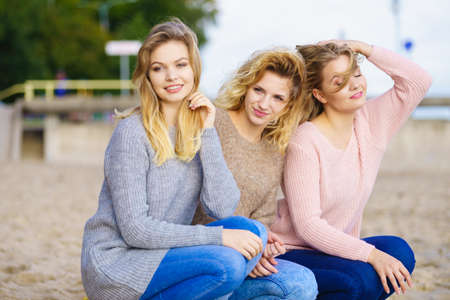 Three fashionable women wearing sweaters during warm autumnal weather spending their free time on sunny beach. Fashion models outdoorの写真素材