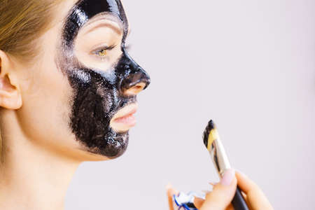 Young woman applying with brush black detox mud mask to her face. Girl taking care of skin. Spa treatment. Skincare. Side viewの写真素材