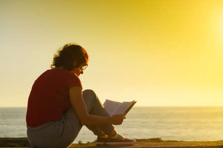 Mature tourist woman relaxing on coast with book, enjoy fresh air in sunset time. Reading on holidays. Relax time.の写真素材