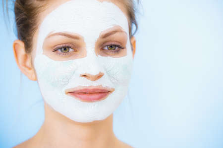 Young woman with white dry mud mask on face, against blue. Teen girl taking care of oily skin, cleaning the pores. Beauty treatment. Skincare.の写真素材