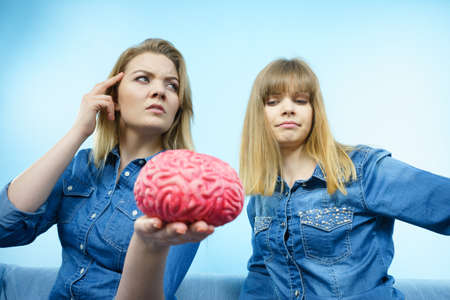 Friendship, human relations concept. Two women friends or sisters wearing jeans shirts, thinking about solving problem holding fake brain.の写真素材