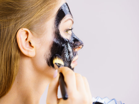 Young woman applying with brush black detox mud mask to her face. Girl taking care of skin. Spa...